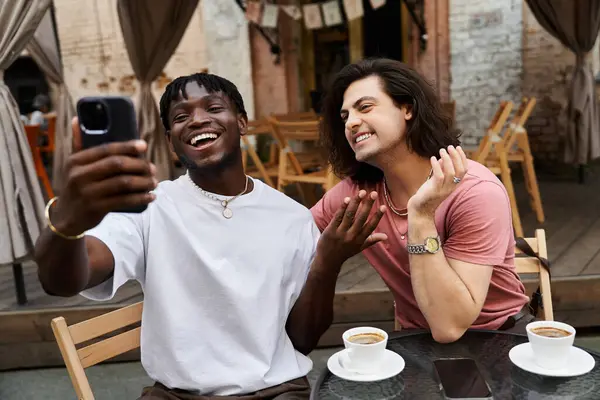 Two handsome men laugh together while taking a selfie during their romantic cafe date. — Stock Photo