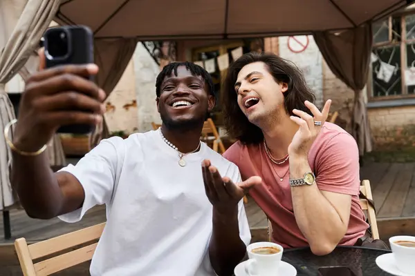 A loving gay couple laughs together at a cozy cafe, capturing memories on their phones. — Stock Photo