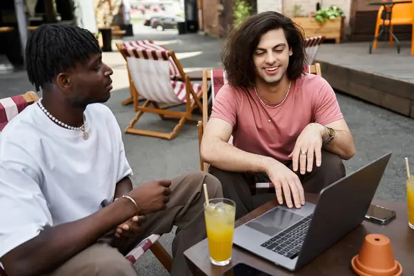 Two handsome men share smiles and a laptop, sipping bright drinks under the warm sun at a cafe. — Stock Photo