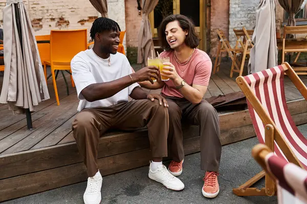 Two handsome men share laughter and toast with their drinks while sitting at a lovely cafe terrace. — Stock Photo