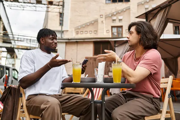 Dois homens bonitos estão compartilhando risos e bebidas em um café encantador durante uma tarde ensolarada. — Fotografia de Stock