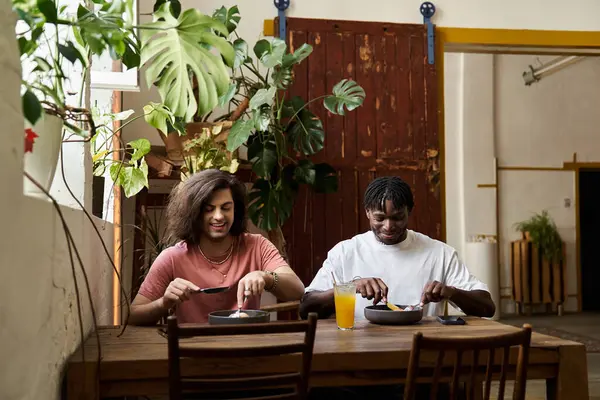 Due uomini belli sedersi da vicino a un tavolo di legno rustico in un caffè tranquillo, condividendo il pranzo e sorrisi. — Foto stock