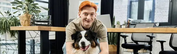 Un giovane uomo dai capelli rossi sorride mentre gioca con un cane in un ambiente ufficio contemporaneo. — Foto stock