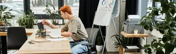 Молодий чоловік з рудим волоссям зосереджується на роботі в стильному офісному столі, оточеному рослинами. — стокове фото