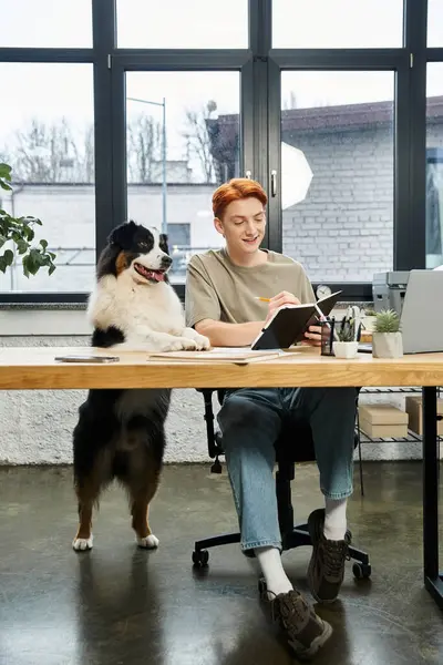 A young man with red hair sits at a desk writing, with a friendly dog by his side. — Stock Photo