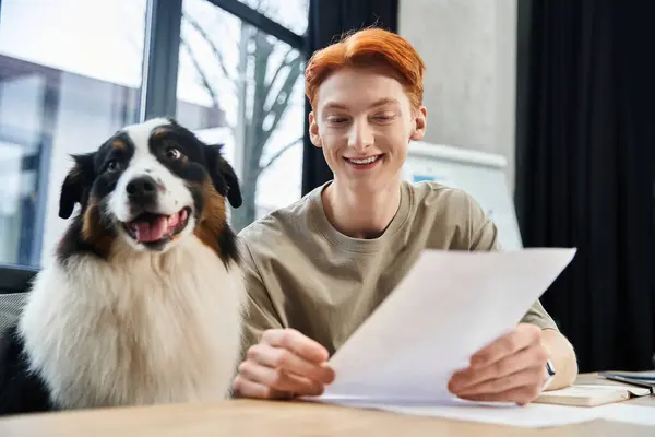Um jovem homem ruivo sorri enquanto revisa documentos com seu cachorro ao seu lado no escritório. — Fotografia de Stock
