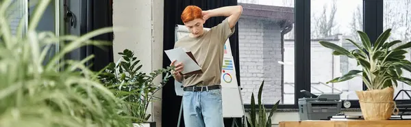 Young red haired man examines paperwork in an office filled with plants and natural light. — Stock Photo