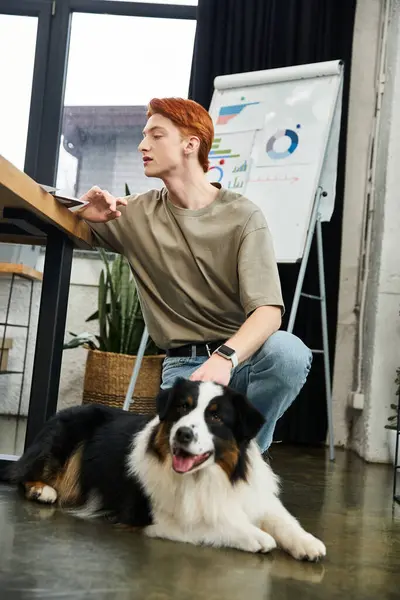 Ein junger Mann mit roten Haaren arbeitet in einem modernen Büro, während sein Hund neben ihm entspannen kann. — Stockfoto