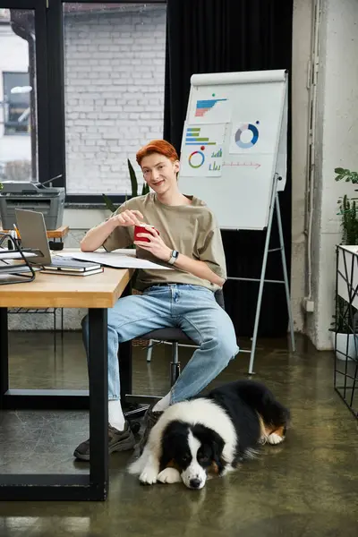 A young man with red hair relaxes at his desk in a lively office with his dog beside him. — Stock Photo