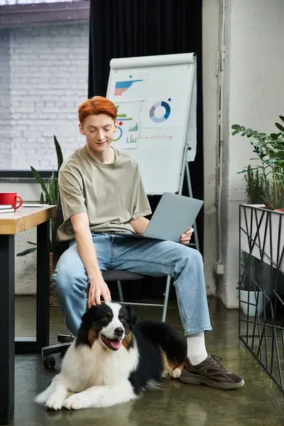 A dedicated young man with red hair interacts with his dog while working in a contemporary office. — Stock Photo