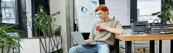 A dedicated young man with red hair focuses on tasks while enjoying a drink in a stylish workspace. — Stock Photo