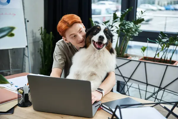 Dedicado joven con el pelo rojo se dedica a tareas de trabajo, mientras que su perro amigable se sienta felizmente. - foto de stock