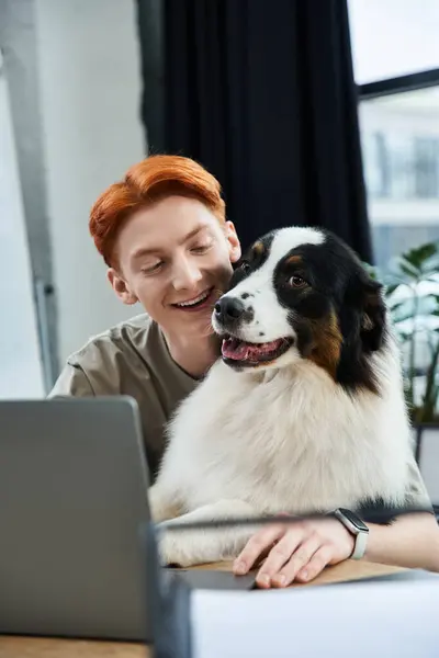 In a contemporary office, a young man with striking red hair smiles while interacting with his dog. — Stock Photo