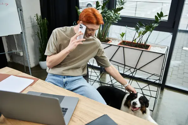 In a contemporary office, a young red haired man enjoys a call while petting his dog by his side. — Stock Photo