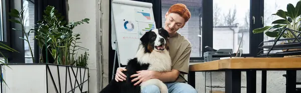 A dedicated young man with red hair takes a break at work, happily interacting with a large dog. — Stock Photo