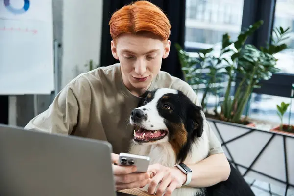 In a modern office, a young man with red hair checks his smartphone while a dog sits nearby. — Stock Photo