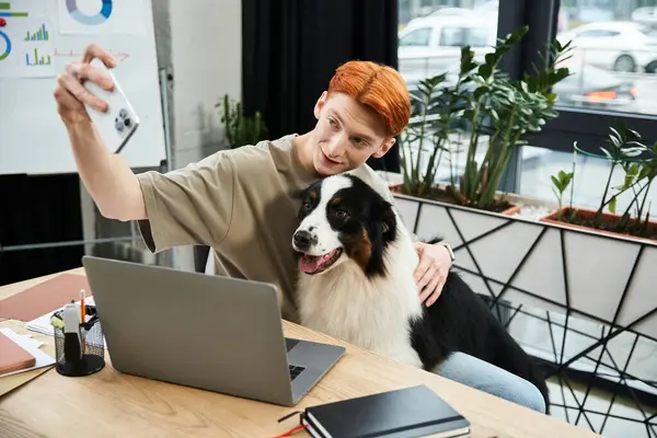 Young man with vibrant red hair happily takes a selfie with his dog while working at a desk. — Stock Photo