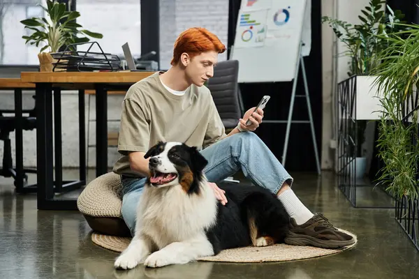 Focused young man checks his phone while sitting on the floor with his dog in a stylish office. — Stock Photo