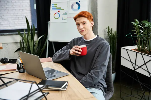 Red haired man smiles as he takes a break with a coffee cup while working at his desk. — Stock Photo