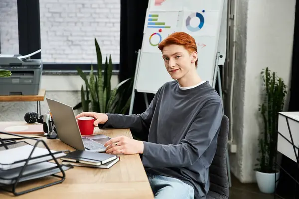 Un joven con el pelo rojo interactúa activamente con su portátil en una configuración de oficina contemporánea. - foto de stock