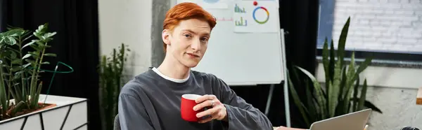 A young man with red hair smiles, holding a coffee cup while focused on his work. — Stock Photo