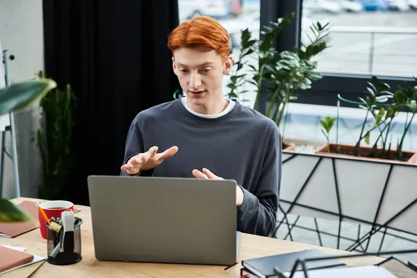 Un giovane uomo dai capelli rossi si impegna in una riunione virtuale alla sua scrivania in un ufficio moderno. — Foto stock