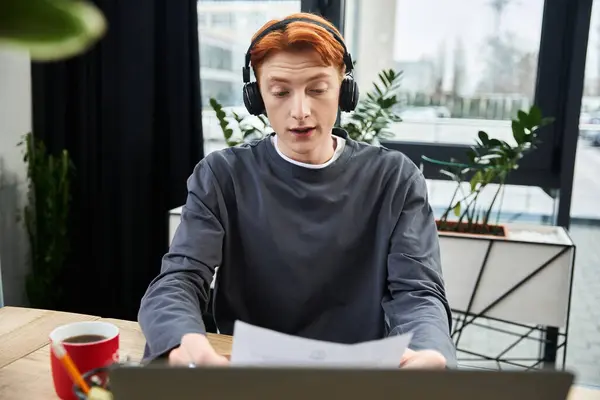 A young man with red hair focuses on his tasks, wearing headphones in a modern workspace. — Stock Photo