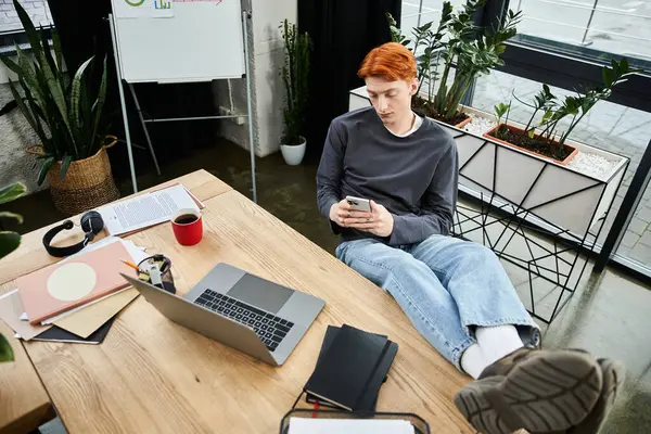 Focused young man with red hair engages with his smartphone amid office tasks and greenery. — Stock Photo