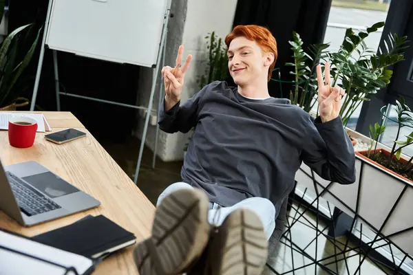 A young red haired man takes a break in a bright office, sitting casually and smiling at his desk. — Stock Photo