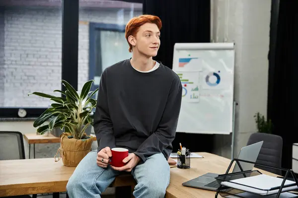 A young red haired man sits casually on a desk, savoring coffee while engaging in work tasks. — Stock Photo