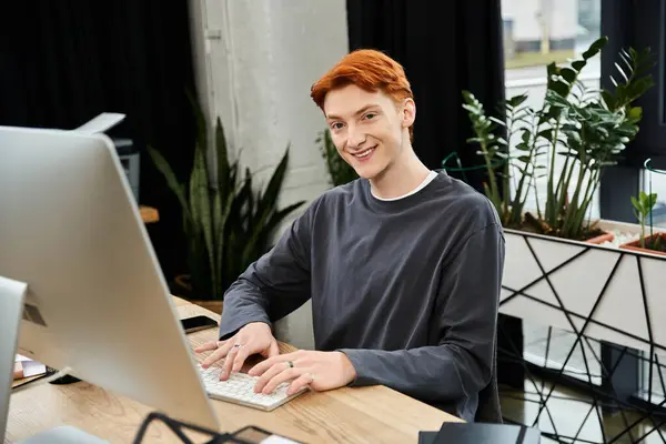 In a bright workspace, a young man with red hair smiles while typing on his computer keyboard. — Stock Photo