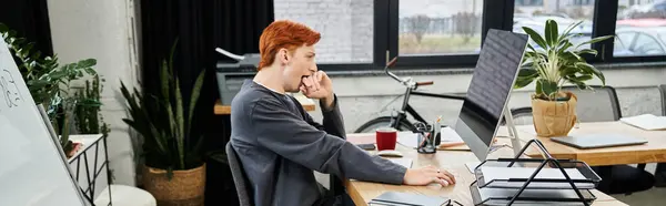 A young red haired man intently works in a modern office filled with greenery. — Stock Photo