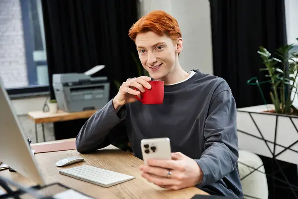 L'uomo dai capelli rossi sorride mentre sorseggia un sorso da una tazza di caffè mentre lavora ai suoi compiti. — Foto stock