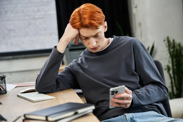 Un giovane uomo dai capelli rossi si siede a una scrivania, contemplando le attività di lavoro mentre guarda il suo telefono. — Foto stock