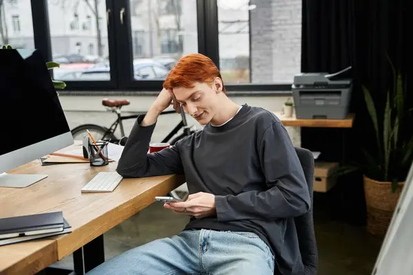Un joven con el pelo rojo se centra en su smartphone en un escritorio de madera en una oficina moderna. — Stock Photo