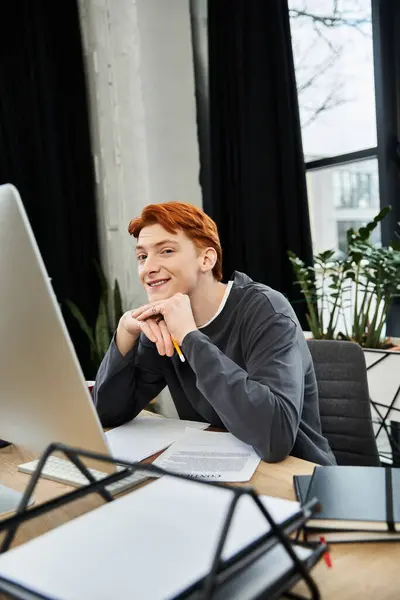 A young man with ginger hair smiles while working at his modern office desk. — Stock Photo