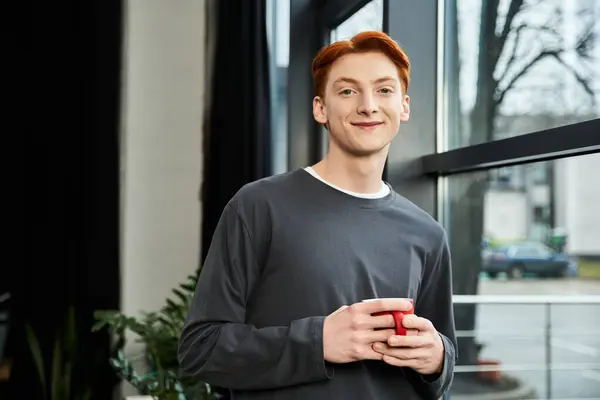 Guapo joven con el pelo rojo se relaja con una taza caliente, mostrando su dedicación en el trabajo. - foto de stock