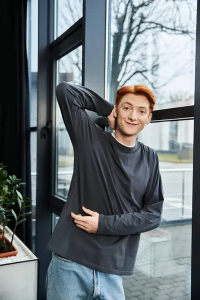 Young red haired man smiles while confidently posing in a bright office space with large windows. — Stock Photo