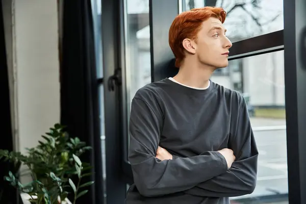 A young man with red hair stands by the window in a modern office, deep in thought. — Stock Photo