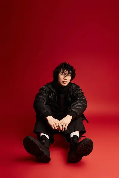Young asian man in stylish black outfit sits on red studio backdrop, exuding cool and confidence. — Stock Photo