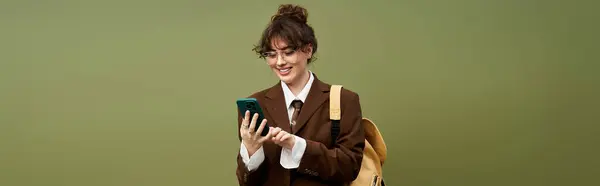 A young woman in a stylish outfit smiles while using her smartphone against a green backdrop. — Stock Photo