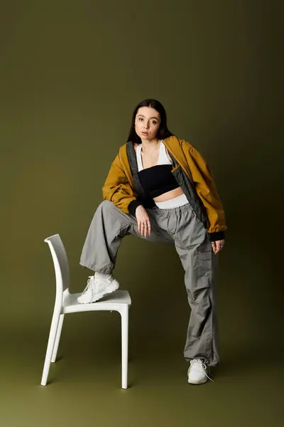 Confident young woman poses creatively with one leg on a white chair, highlighting modern fashion. — Stock Photo
