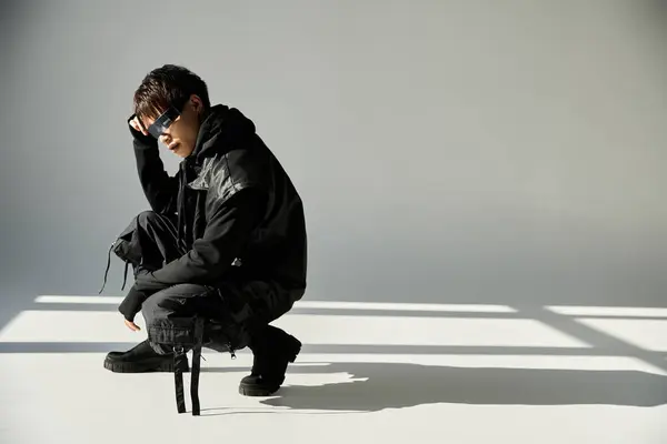 Beau asiatique homme s'accroupit dans un studio moderne, mettant en valeur une tenue élégante sous un éclairage doux. — Photo de stock