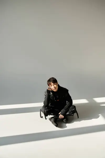 Un beau jeune homme en noir chic s'assoit gracieusement sur le sol, baigné de lumière douce. — Photo de stock