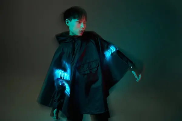 A young asian man in a stylish black poncho showcases vibrant glowing accents in trendy lighting. — Stock Photo