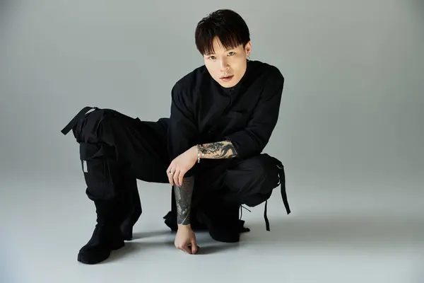 Confident Asian man crouches in a stylish outfit against a neutral backdrop, highlighting fashion. — Stock Photo