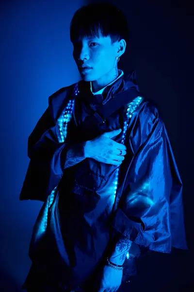 Handsome young asian man poses confidently in striking blue light, dressed in trendy attire. — Stock Photo