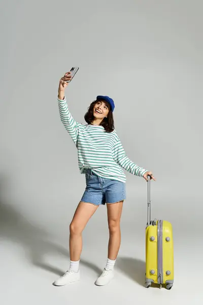 A cheerful young woman in fashionable attire takes a selfie while holding a bright suitcase. — Stock Photo