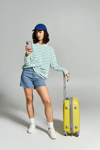 Femme tendance en haut rayé et short en denim pose avec une valise jaune, en utilisant son téléphone. — Photo de stock