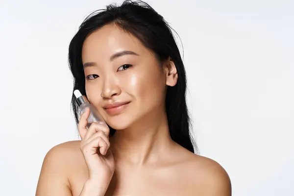 Young Asian woman holds skincare product close to her face, smiling radiantly and confidently. — Stock Photo